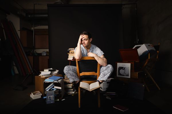 Gregory Nussen sits backwards on a chair, surrounded by stacks of books in a dark room.