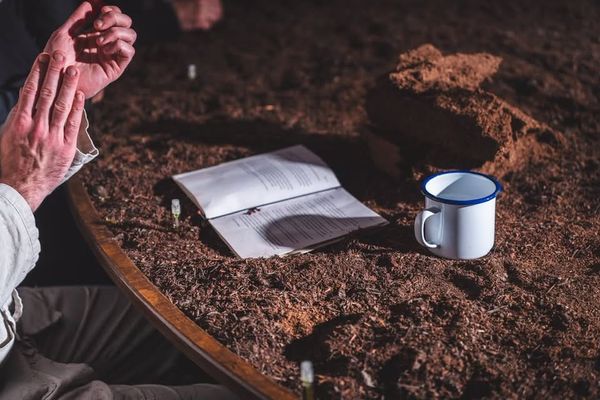 Luke Casserly sits beside a large circular table covered in dark bog soil, his hands raised above an open book and enamel mug.