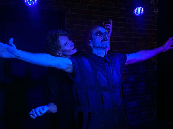 Arthur Angel (Character 1) and Rebecca Howell (Character 2) in Hitherto, arms outstretched under saturated blue stage lighting