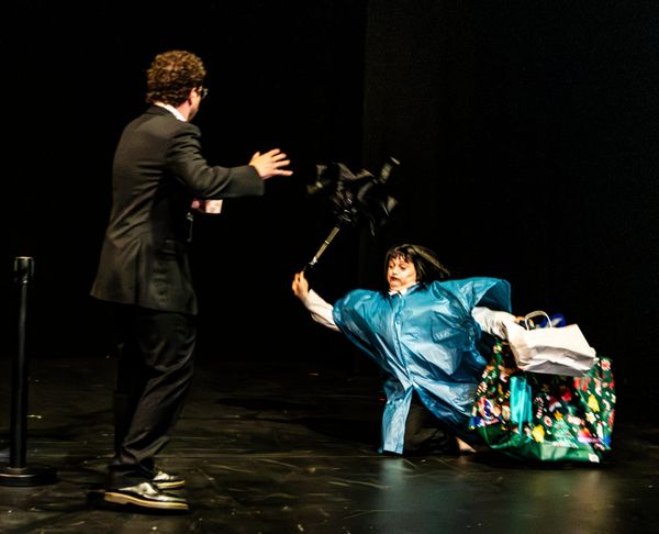 Emilie Bloom, wearing a blue rain poncho and surrounded by colourful bags, tumbles to the floor while holding an umbrella as Tim Sneddon reaches towards her.