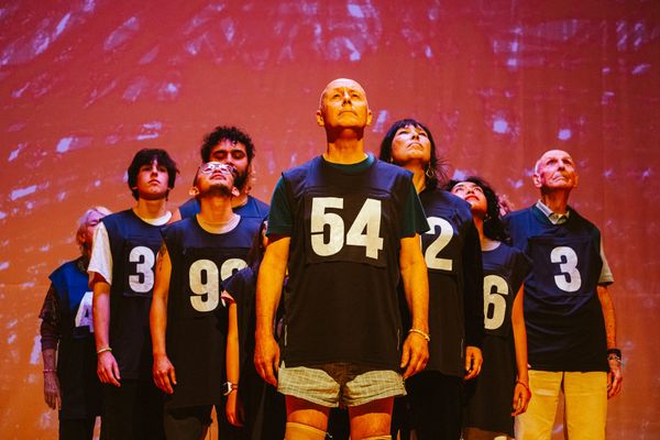 Performers of different generations stand together beneath red lighting, wearing dark shirts with large white numbers that do not correspond to their ages.