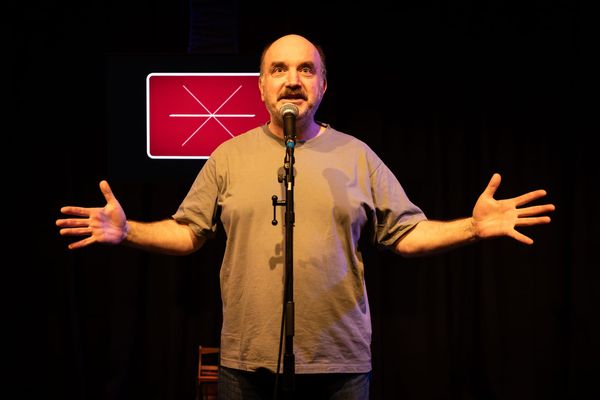Ben Grant stands behind a microphone in Wrong Way Grow Back at La Mama HQ, his arms spread wide. A red geometric image glows on a screen behind him.