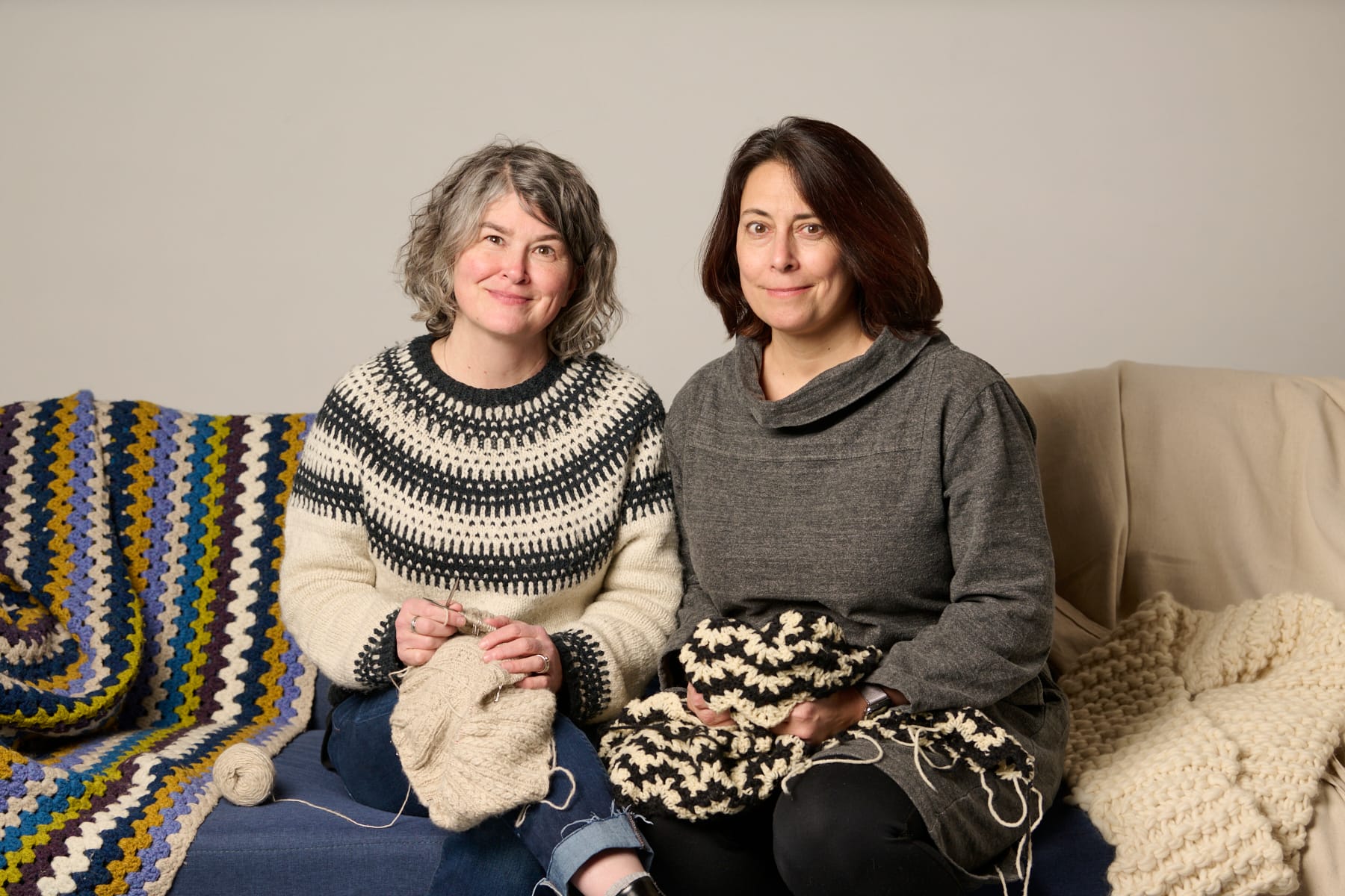 Two women sitting on a couch covered with crocheted blankets. They are each holding a yarn work in progress.