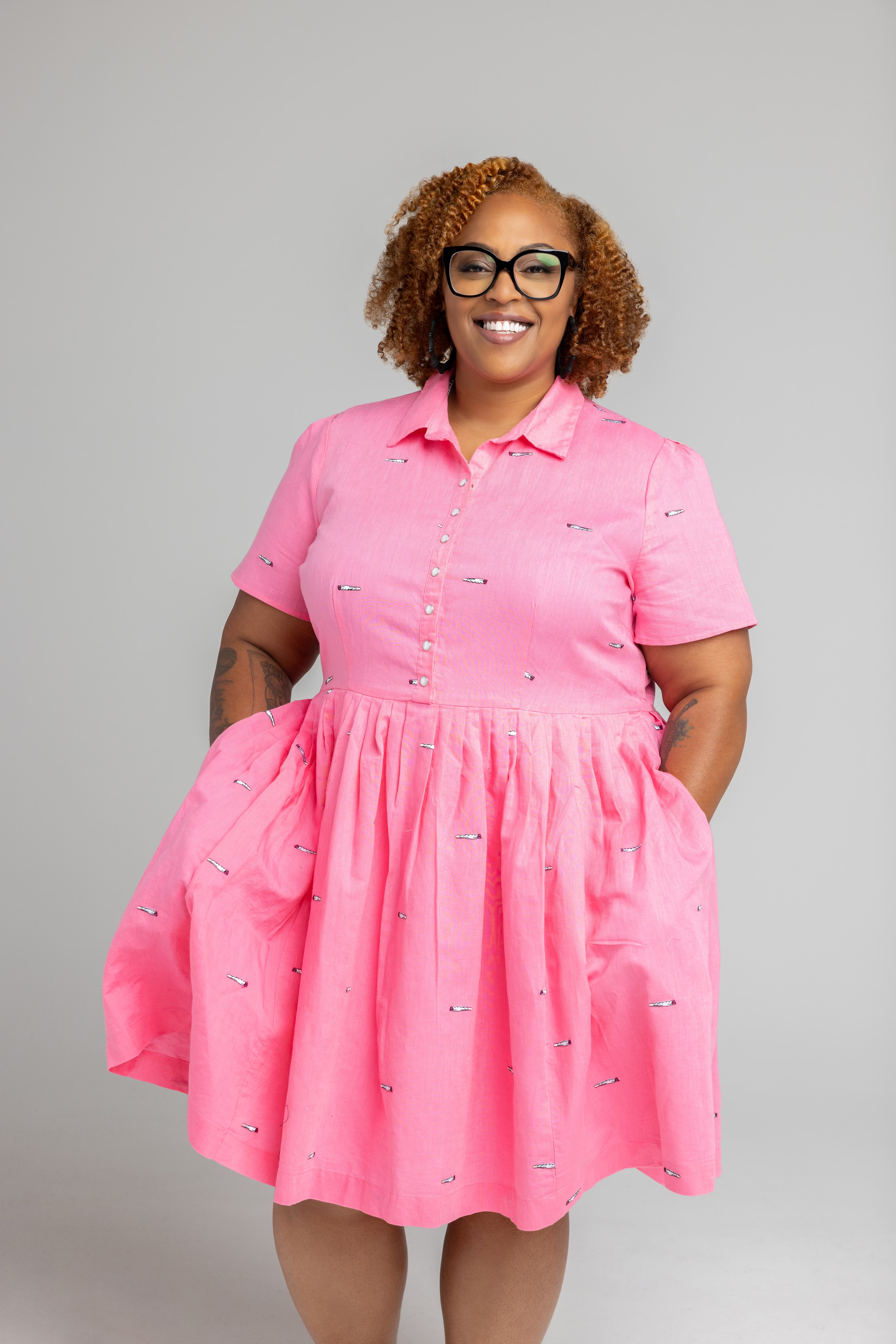 The image shows a person with curly hair wearing glasses and a joyful expression. They are dressed in a bright pink, short-sleeved dress with a collared neckline. The dress features small, embroidered patterns scattered throughout. The dress has a fitted bodice with buttons down the front and a gathered skirt that adds volume. The person's hands are relaxed by their sides, partially in the dress's pockets, standing against a simple, light gray background.