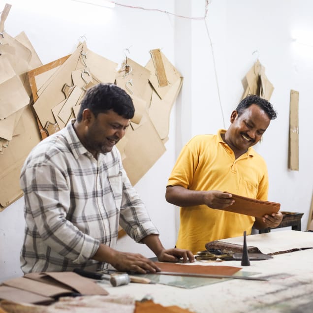 Two men in a workshop smile while working with leather. One wears a plaid shirt, the other a yellow shirt. Pattern templates hang on the wall behind them.