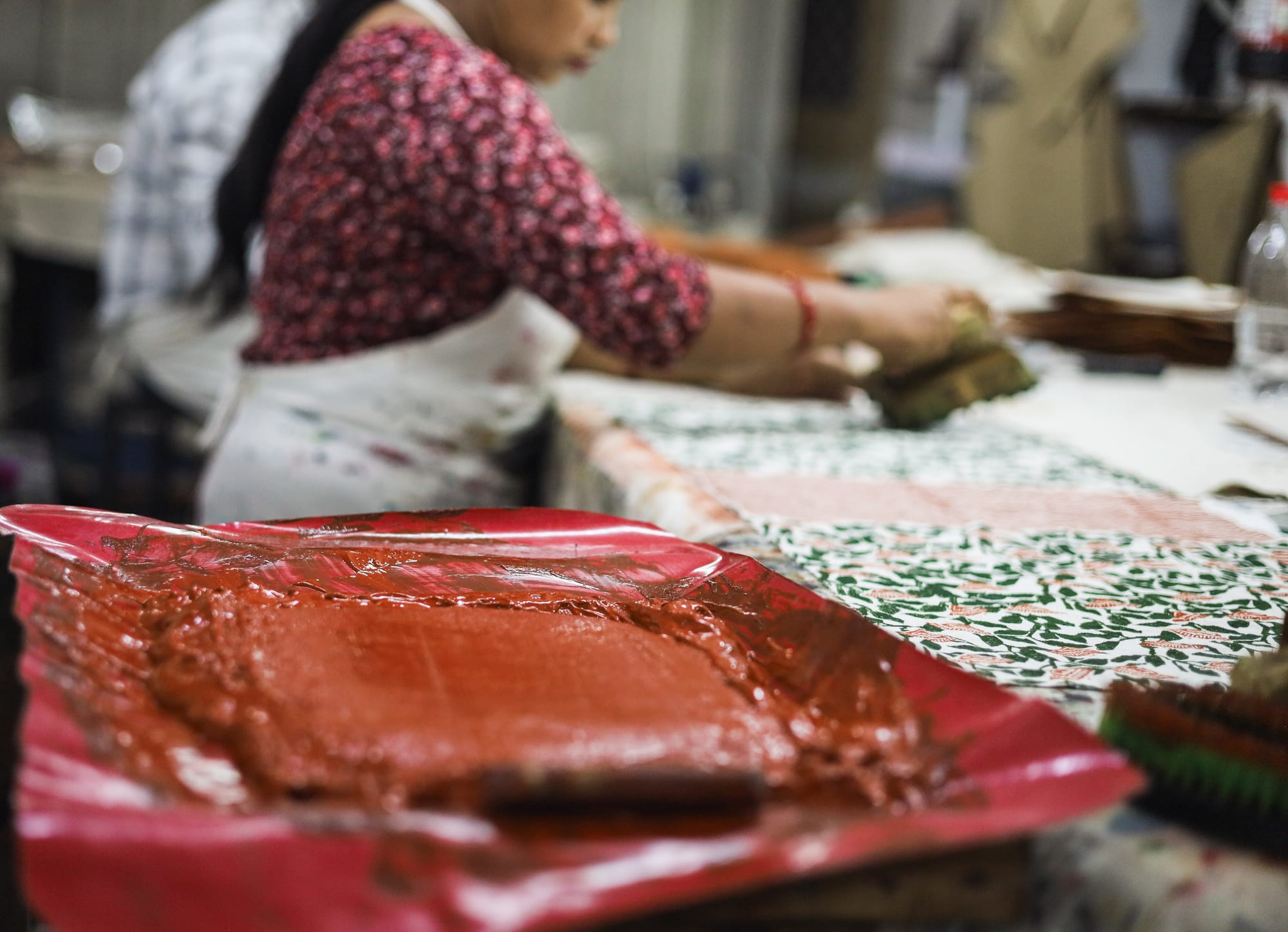 A woman in a patterned red shirt and apron block prints fabric with intricate green designs, using a red dye. The setting is a busy, artistic workspace.