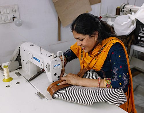 A woman wearing an orange scarf smiles while sewing fabric on a white sewing machine, creating a warm and industrious atmosphere in a workshop.