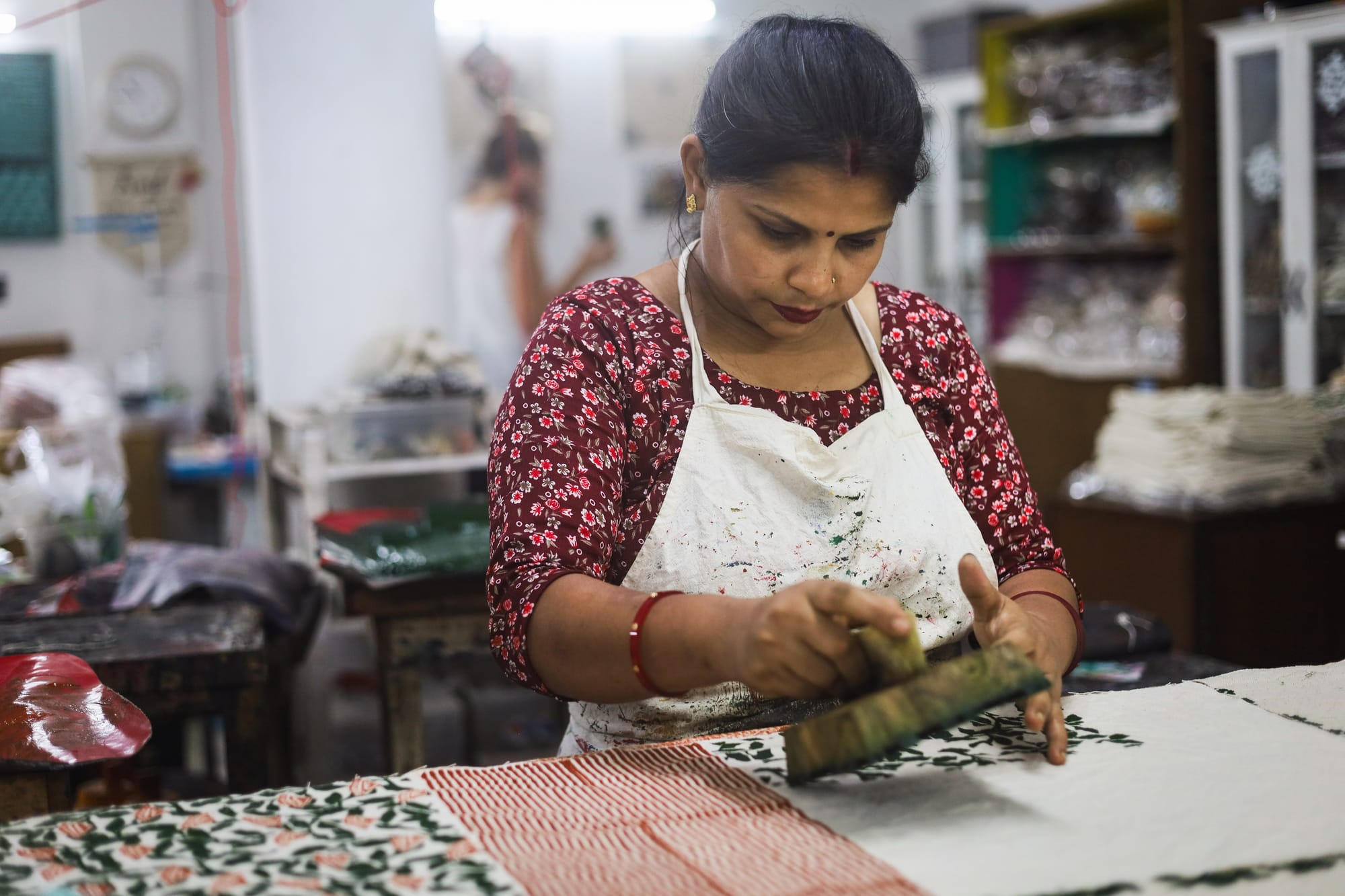 A woman in a patterned red dress focuses intently on block printing fabric in a workshop. Shelves with materials are in the background, creating a busy and creative atmosphere.