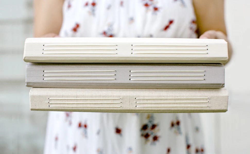 A person in a floral dress holds three closed, beige and gray hardcover books stacked horizontally. The scene conveys a calm, literary feel.