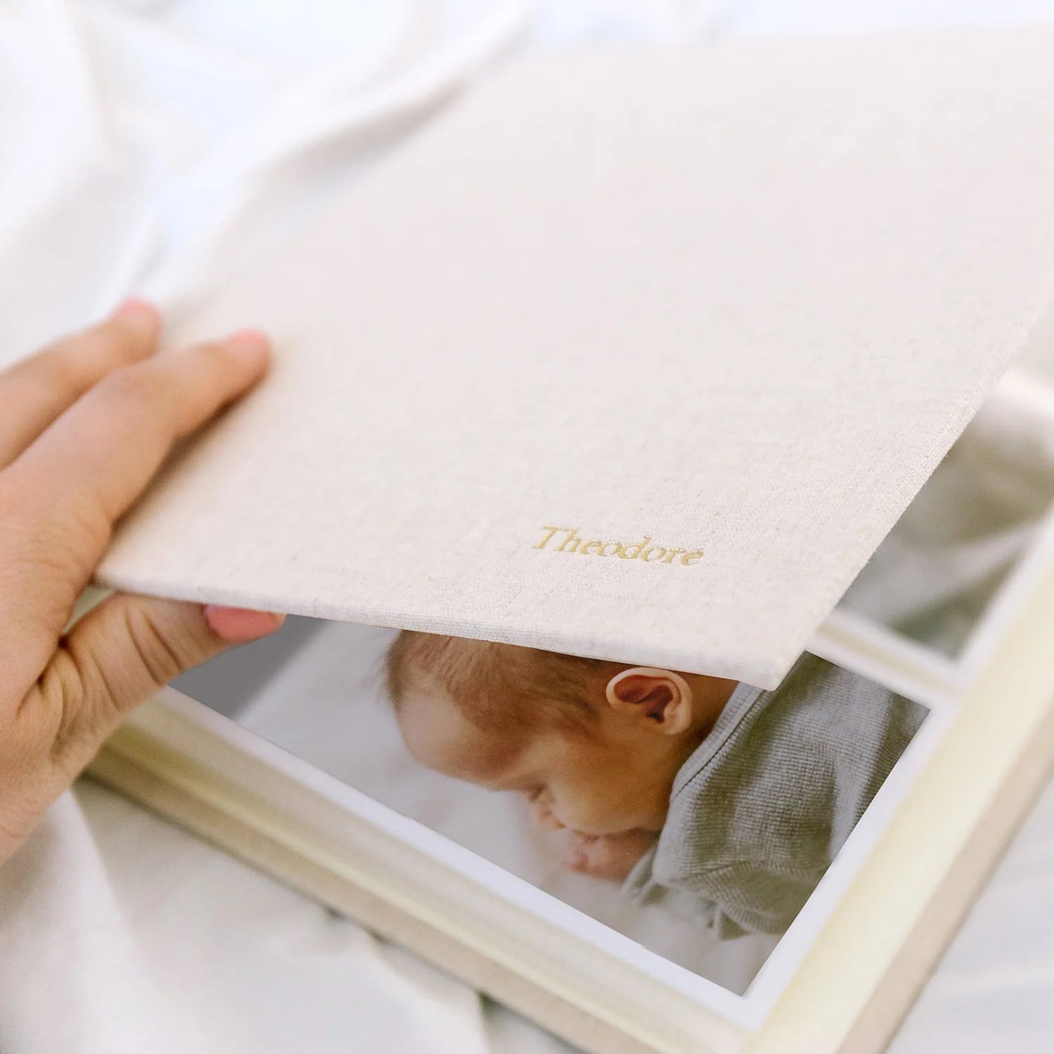 A hand opens a cream-colored photo album with "Theodore" embossed in gold. Inside, a photo of a sleeping baby is visible. The scene feels warm and nostalgic.