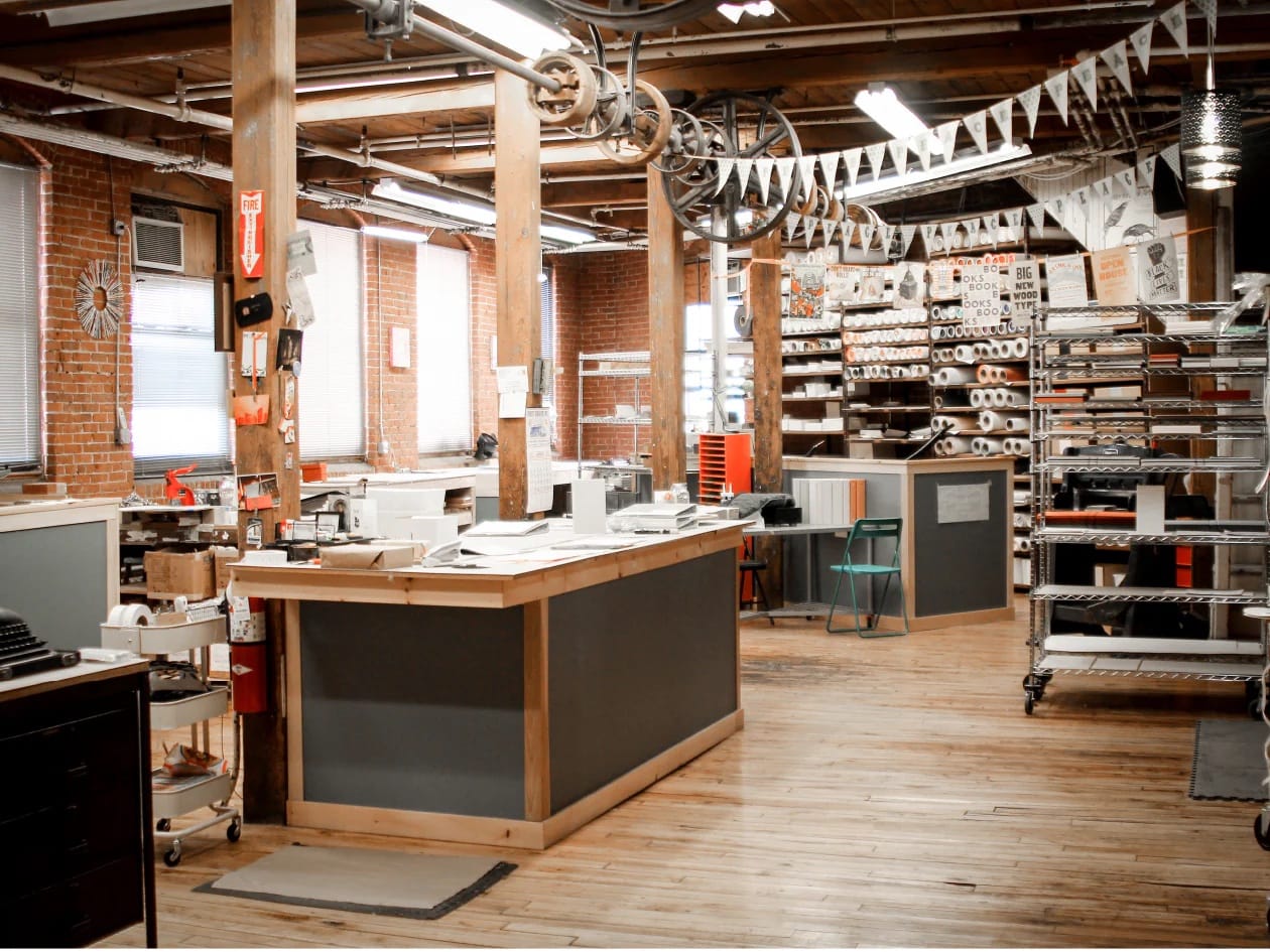 Spacious, slightly rustic office with wooden floors and exposed brick walls. Papers and office supplies on desks, shelves filled with neatly organized rolls.