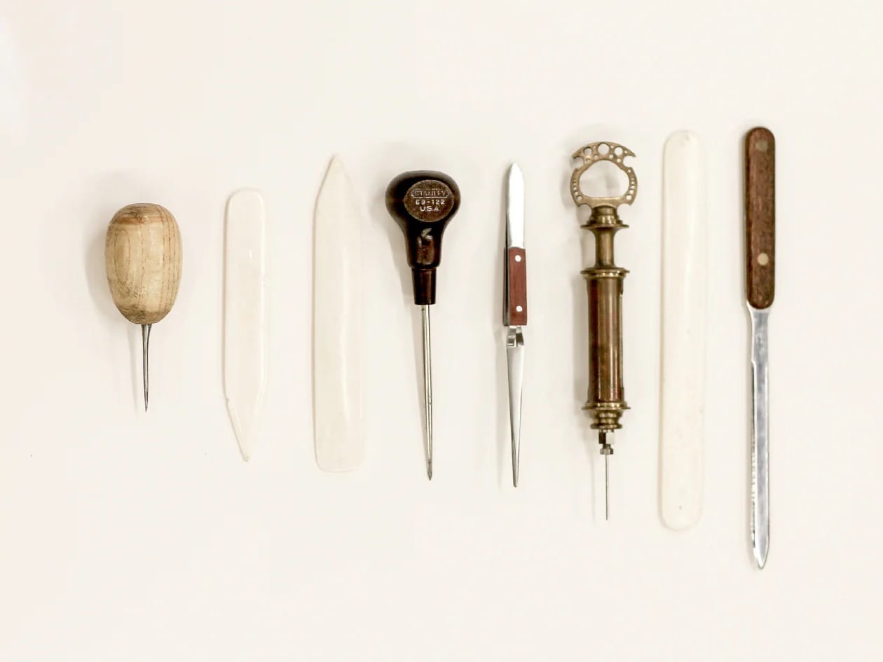 A collection of vintage bookbinding tools displayed on a white background. Includes various awls, bone folders, and knives, evoking a sense of craftsmanship.