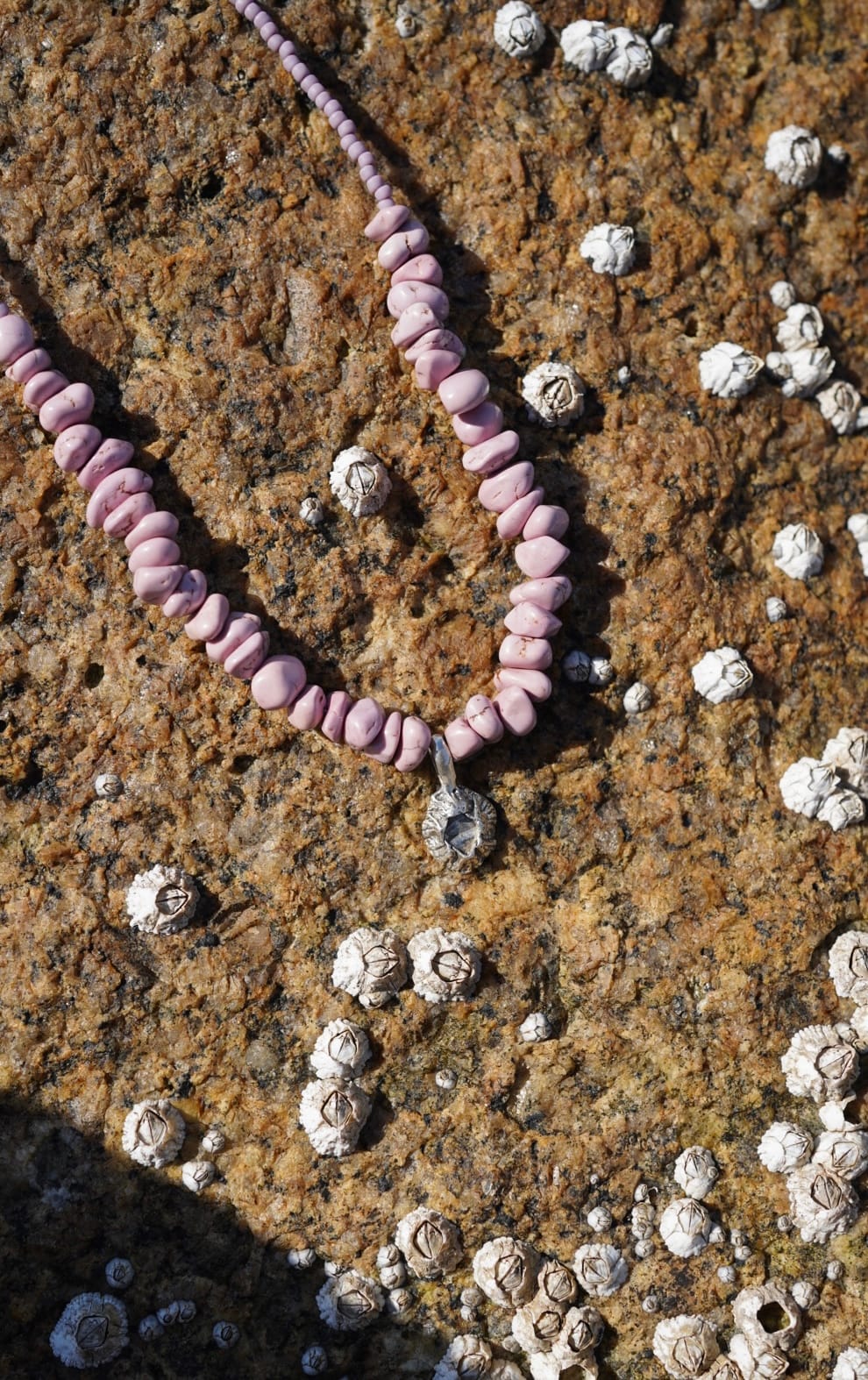 Pink stone necklace with a small pendant lies on a barnacle-covered rock surface. The contrast highlights the necklace's delicate texture and natural tone.