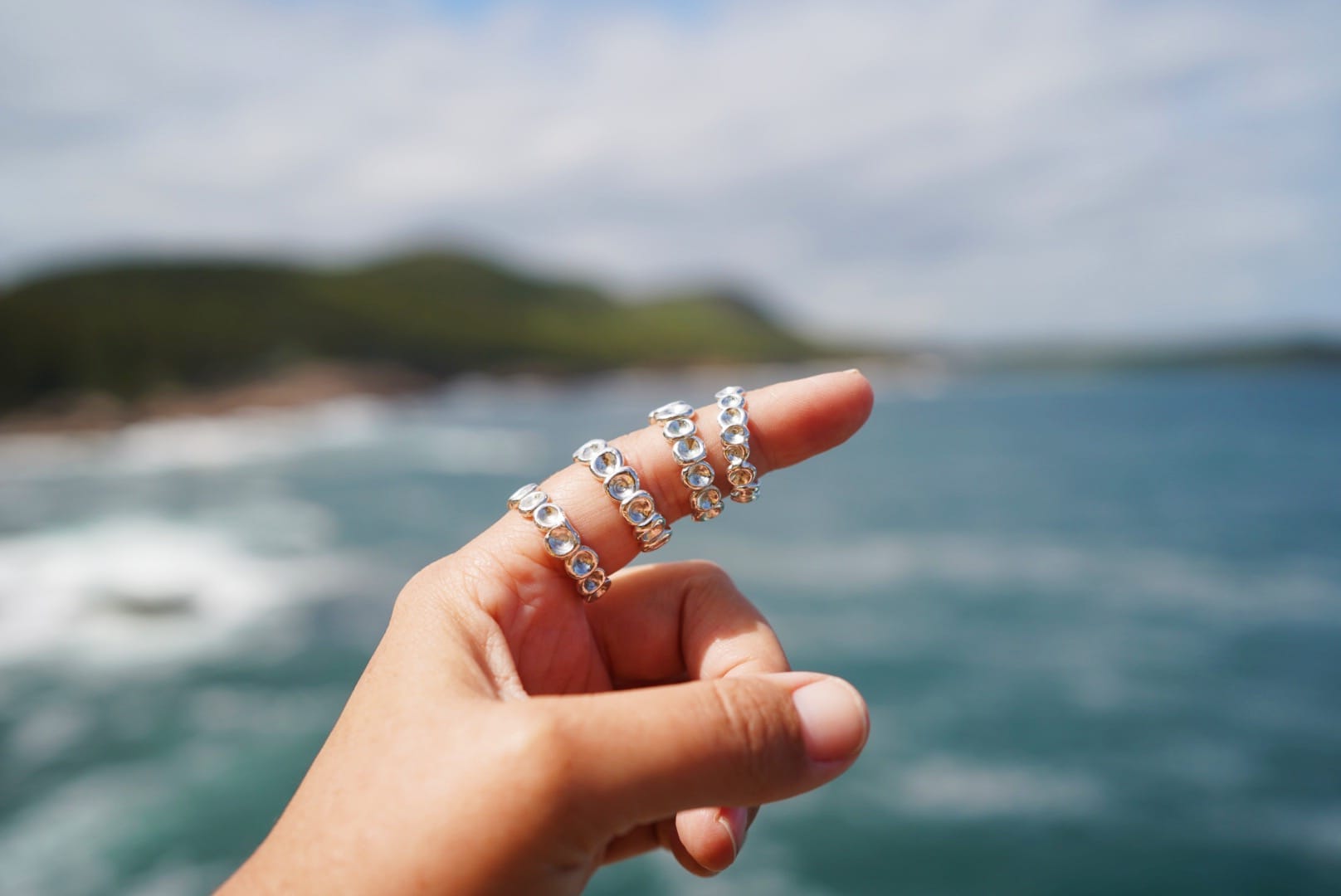 A hand with four silver rings on the thumb is held up against a blurred ocean backdrop. The rings sparkle in the sunlight, conveying elegance.