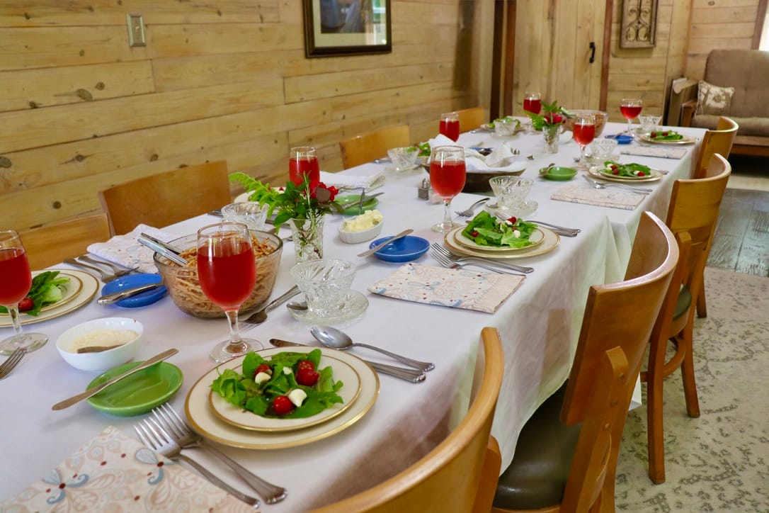 A dining table set for eight with salads, red drinks, and wooden chairs in a rustic room.