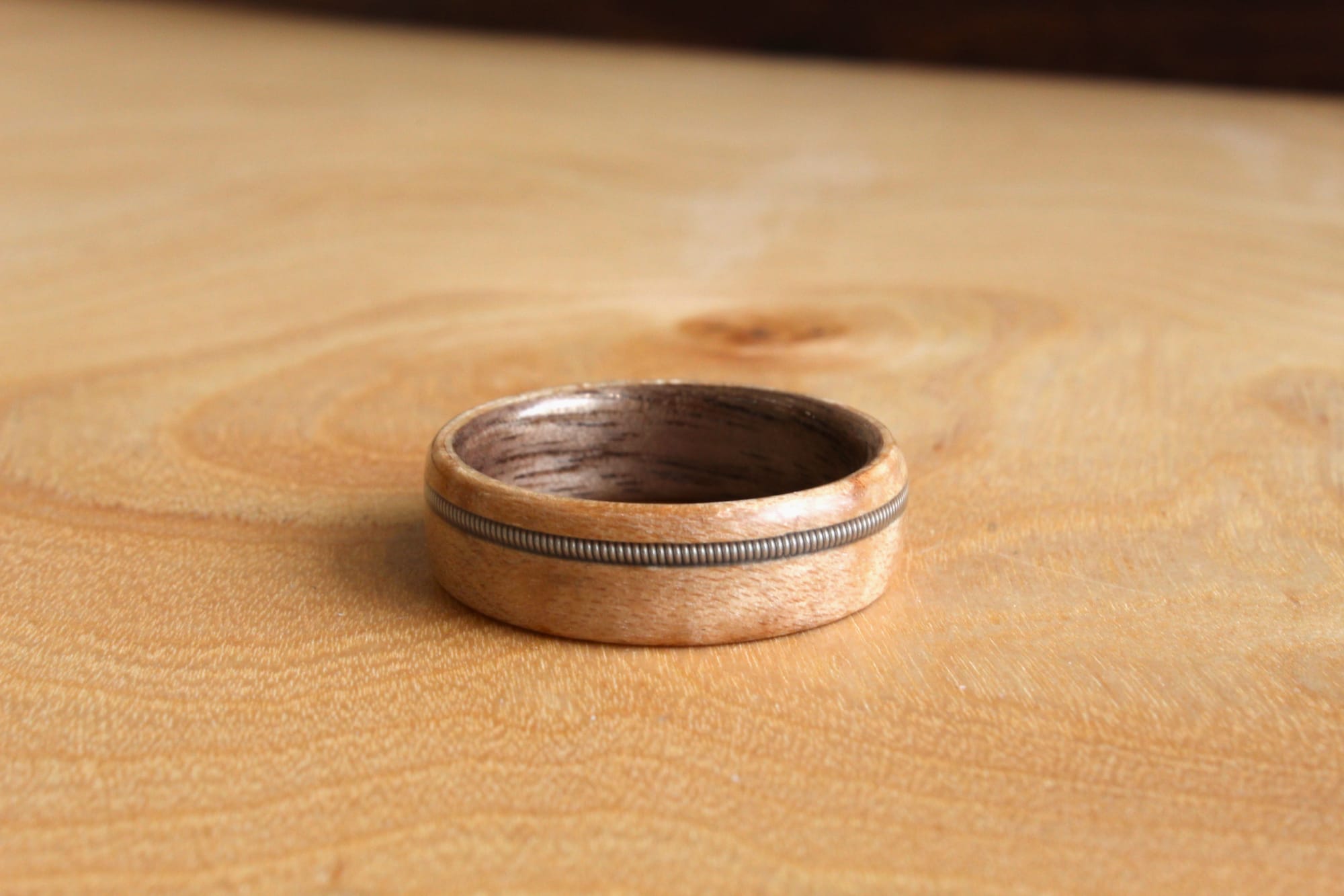 A wooden ring with a smooth finish, featuring a subtle, dark metal inlay around its center made from a bass string, rests on a light wooden surface.