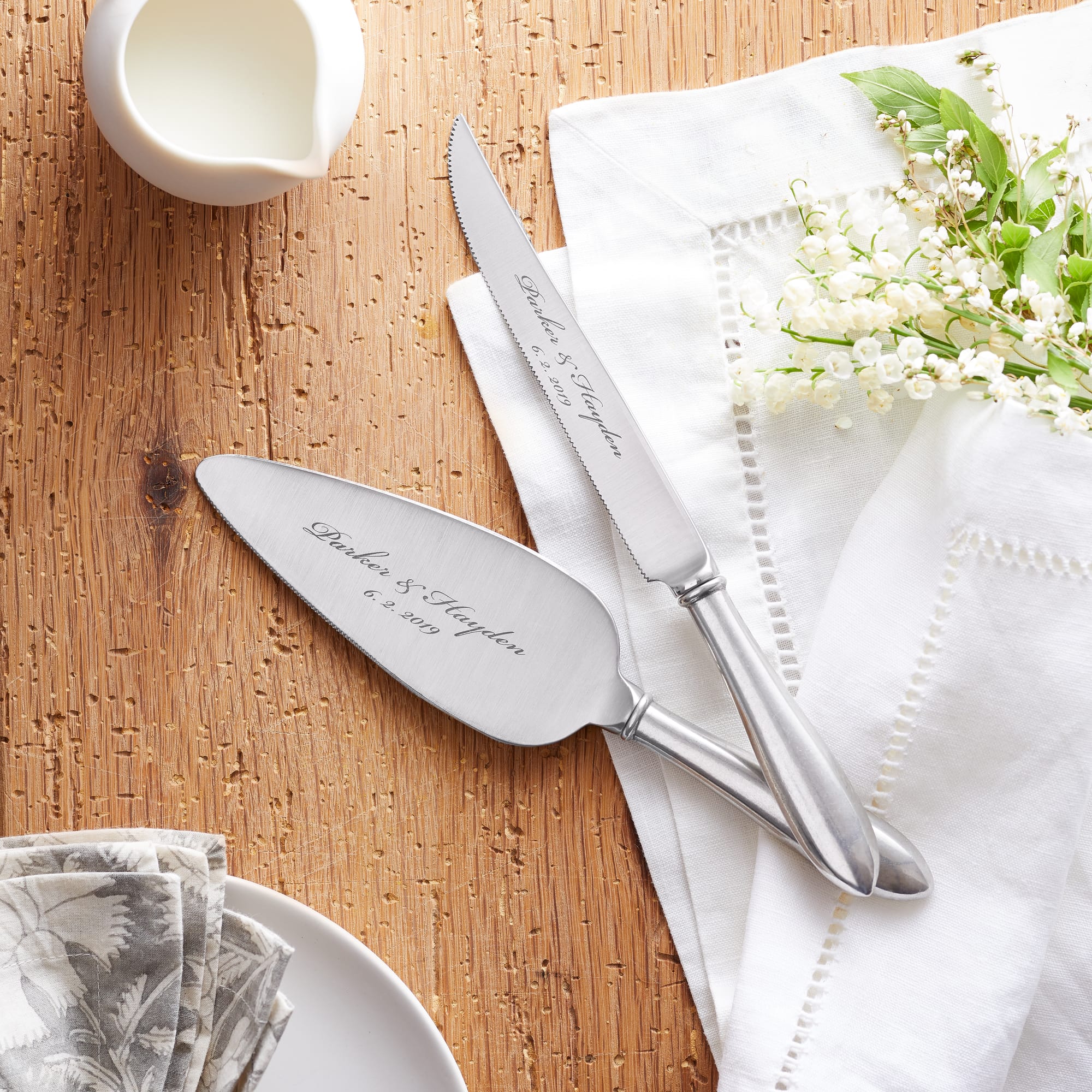 Wedding cake server and knife on a wooden table with white embroidered napkins and lily of the valley flowers, beside a small milk jug. Elegant and celebratory.