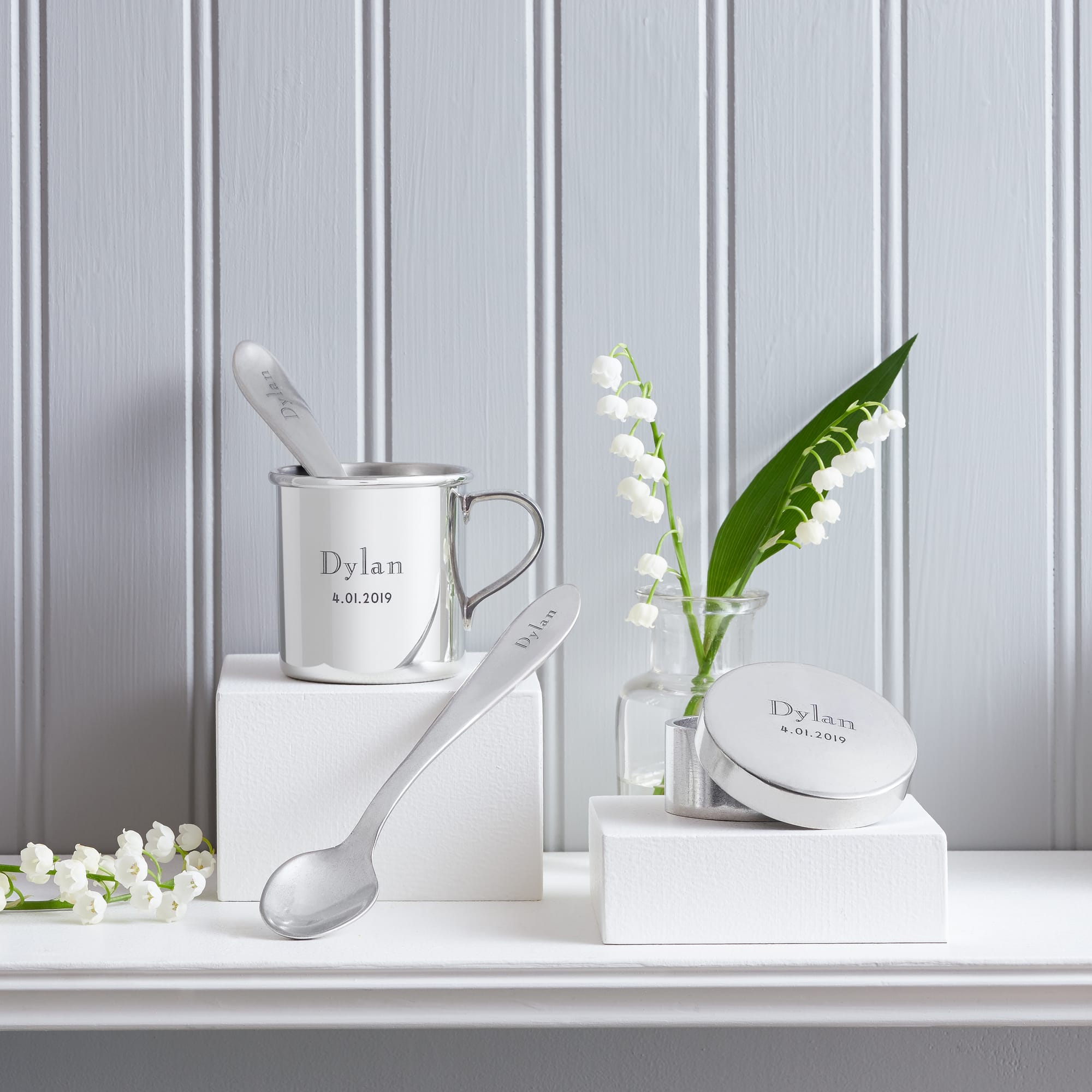 Pewter baby cup, spoon, and keepsake box engraved with "Dylan 8.01.2019" on white pedestals. Lily of the valley flowers add a delicate touch.