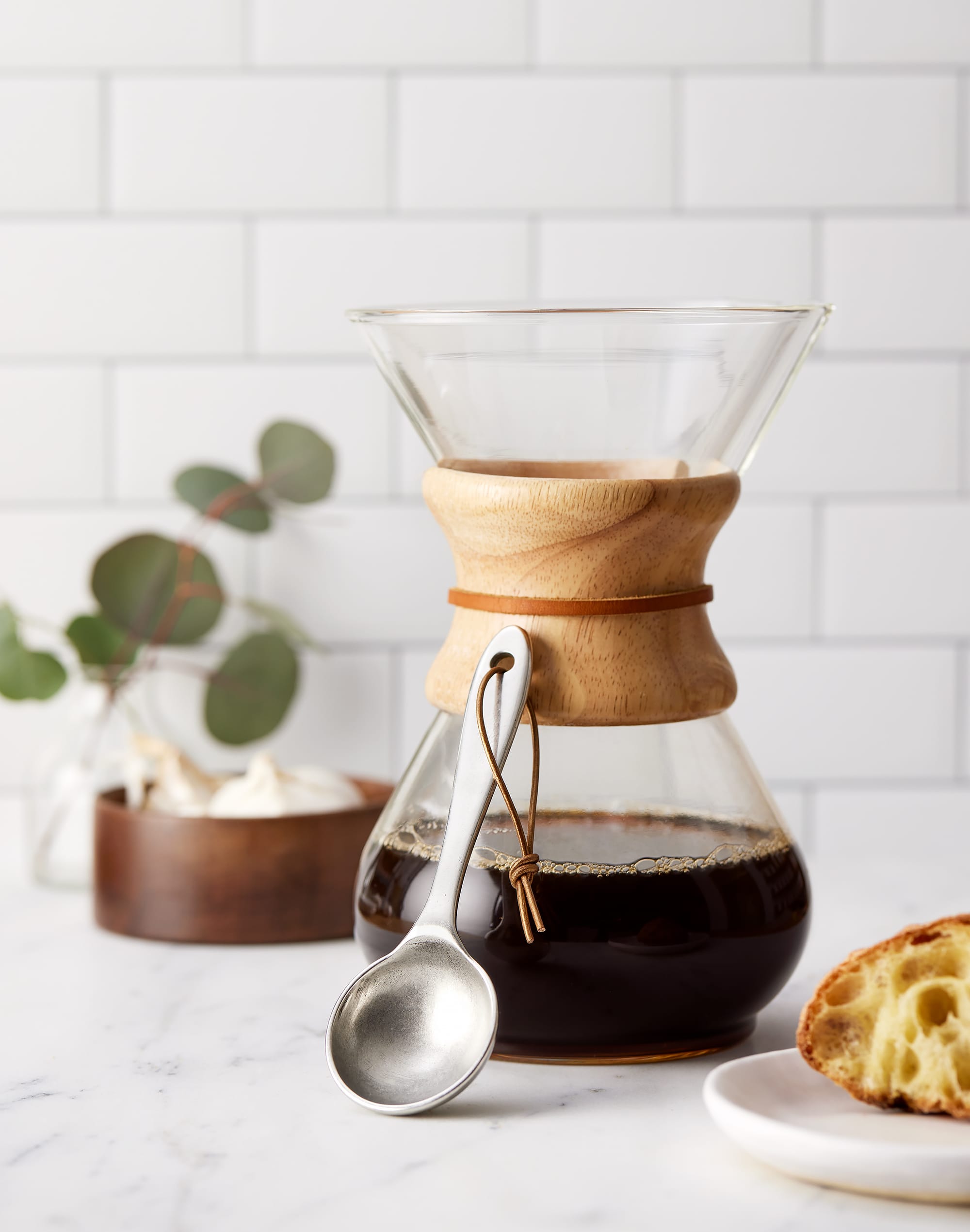 A handcrafted pewter coffee scoop resting vertically on a pour-over glass coffee maker with wood collar.
