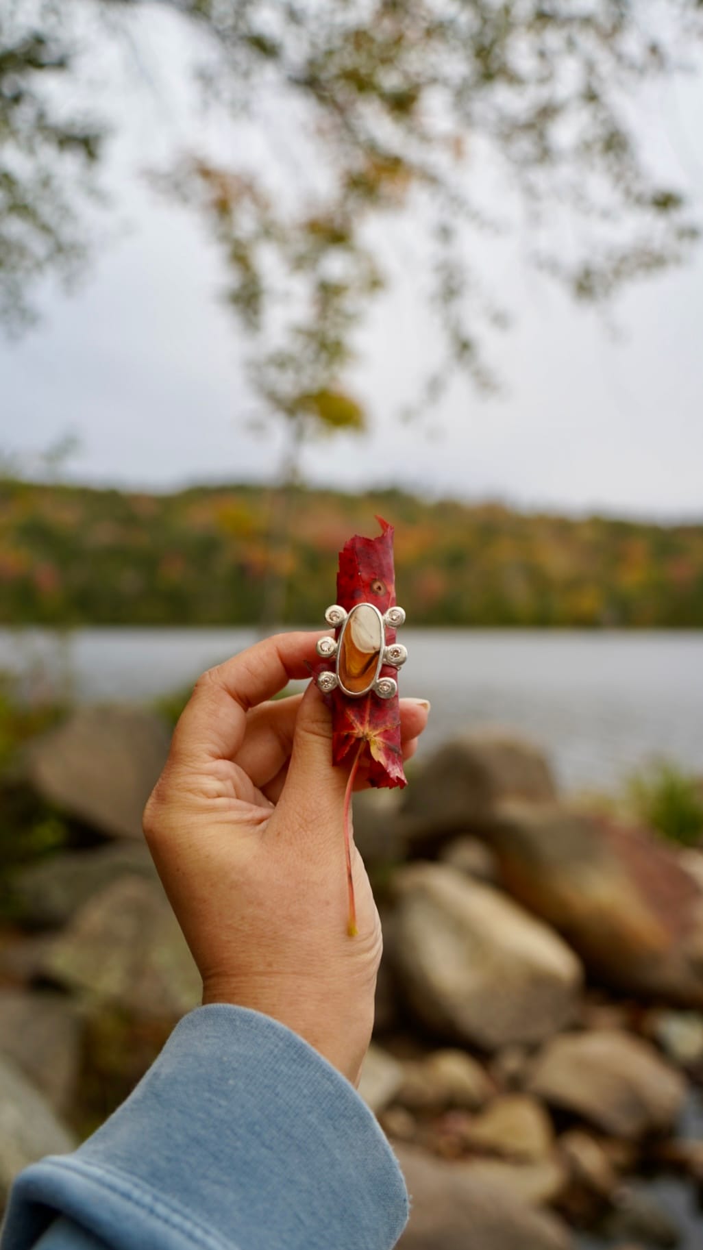 A hand wearing a blue sleeve holds a red leaf adorned with a silver ring in front of a serene lake. Blurred autumn trees and rocks are in the background.