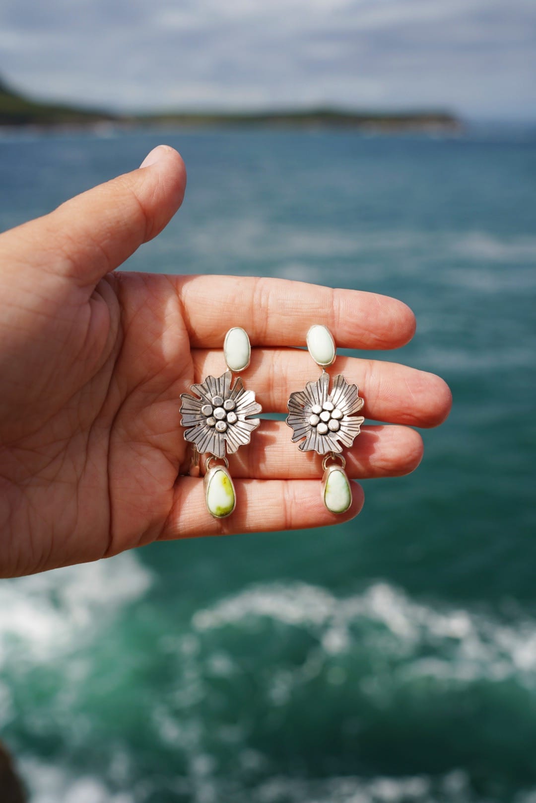 A hand holds silver floral earrings with light green stones against a backdrop of ocean waves, conveying a serene coastal vibe.