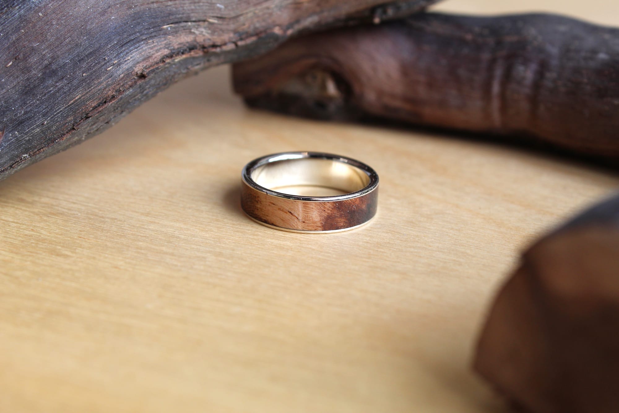 A wooden ring with a polished finish sits on a light wood surface, flanked by smooth, dark driftwood pieces, creating a rustic, natural ambiance.