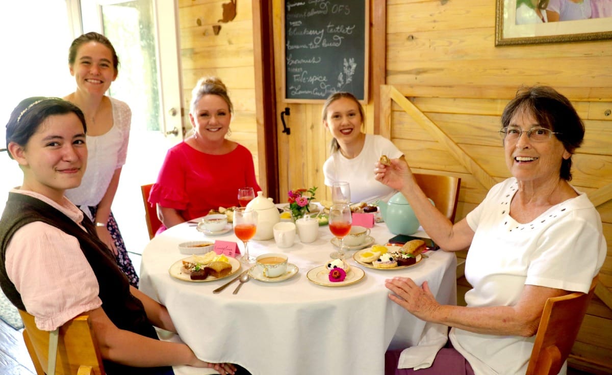 A group of five people smiling around a table set with tea and desserts inside a wooden room.