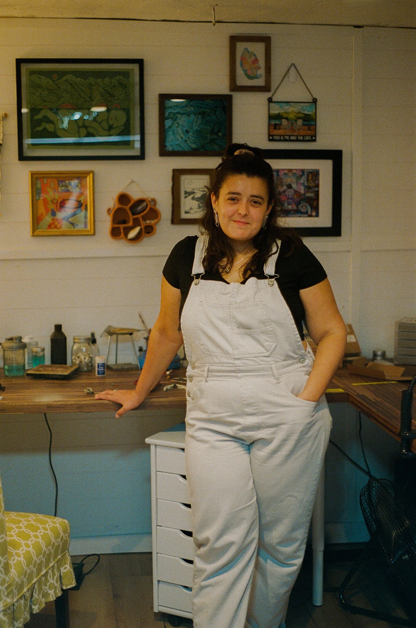 Young woman in white overalls and black shirt, leaning on a workbench in an art studio. Framed artworks are on the wall, creating a creative atmosphere.