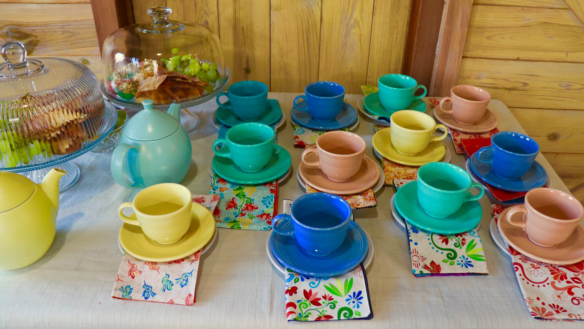 A table with colorful teacups, saucers, patterned napkins, and two ceramic teapots, with pastries and grapes in glass-covered stands.