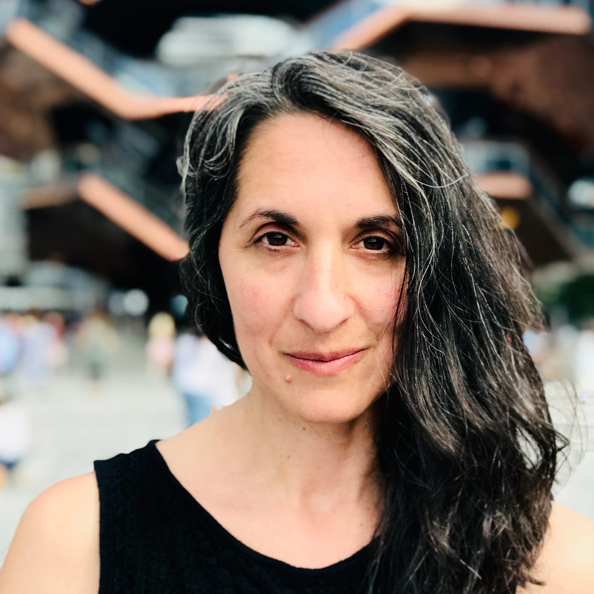 Portrait of a woman with long, gray-streaked dark hair, wearing a black top, standing outdoors with geometric architecture blurred in the background.