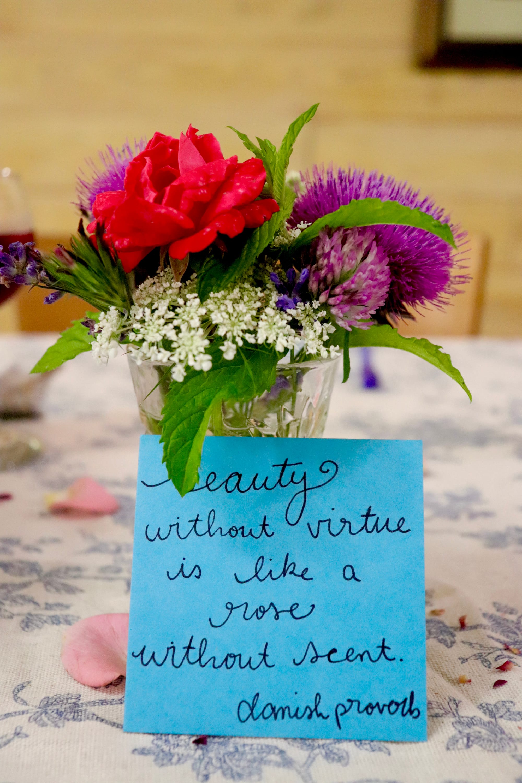 A glass vase with a bouquet of red, purple, and white flowers, accompanied by a blue card with handwritten text.  Transcribed Text:  Beauty without virtue is like a rose without scent. Danish proverb