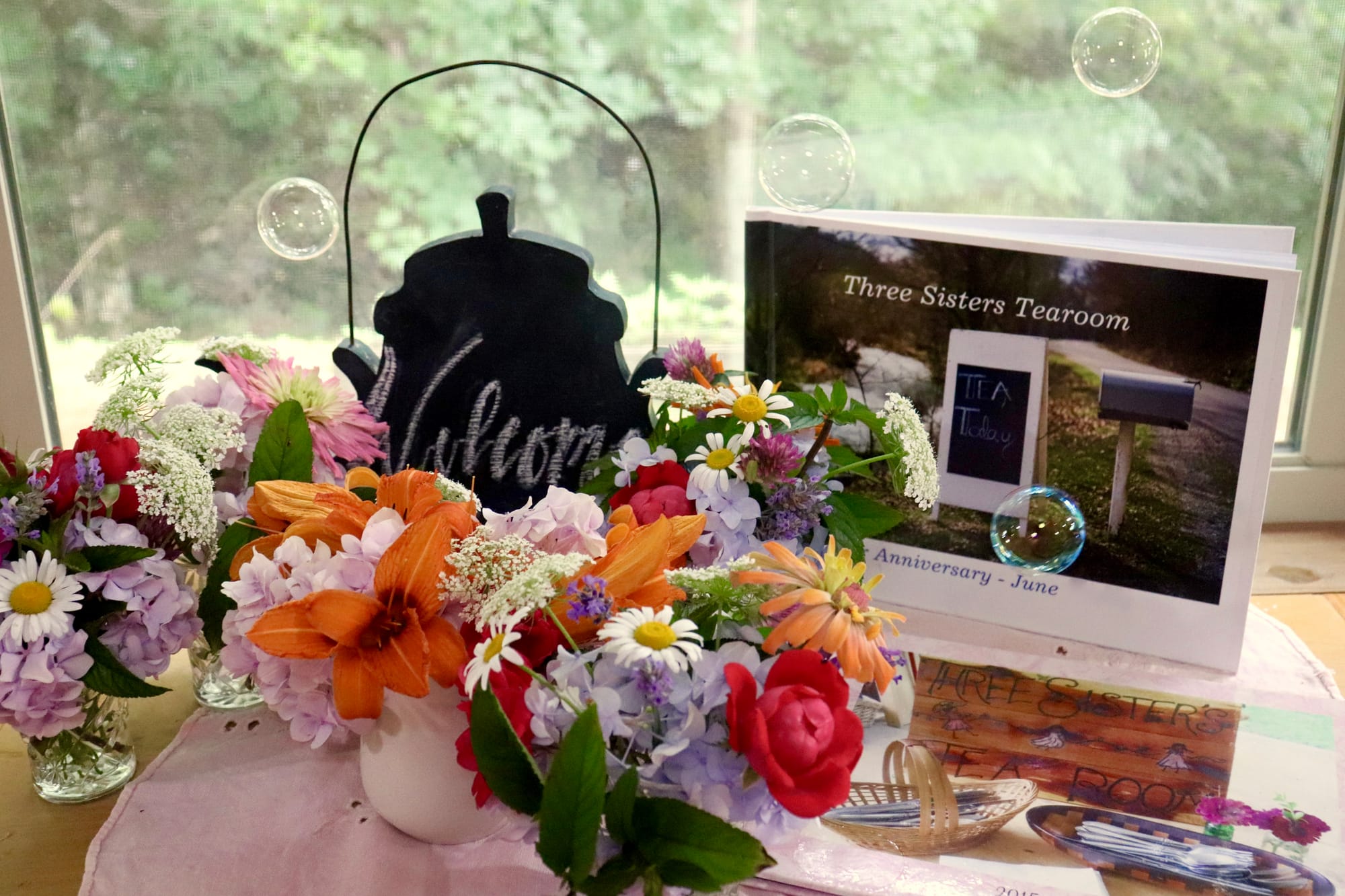 Bouquets of mixed flowers with a welcome sign and a booklet titled "Three Sisters Tearoom" on a table by a window.  Transcribed Text:  Three Sisters Tearoom Tea Today Anniversary - June