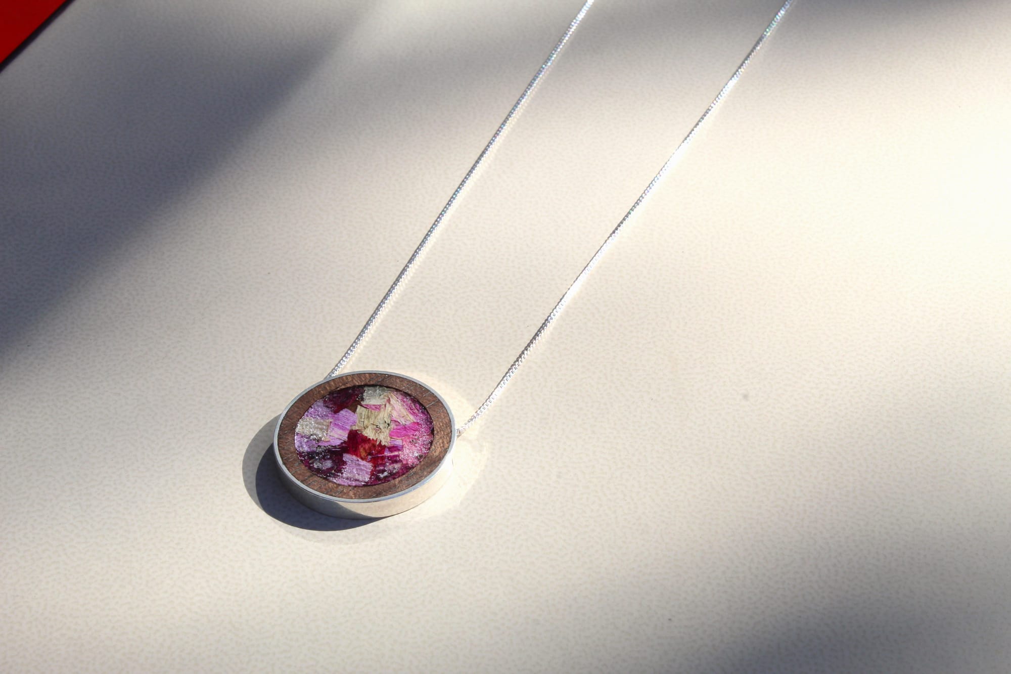 A silver necklace with a circular pendant featuring a vibrant purple and pink abstract pattern rests on a smooth, light surface, lit by soft sunlight.