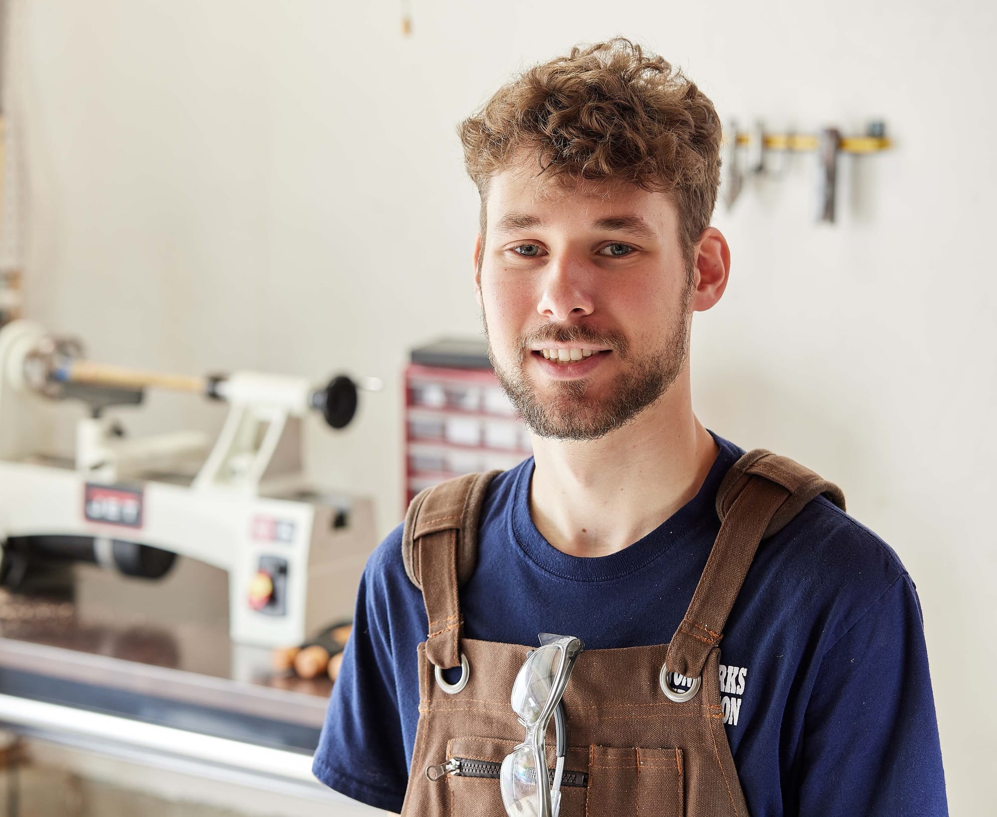 A young man with a beard and curly hair smiles in a workshop, wearing a blue T-shirt and brown overalls. A lathe and tools are visible in the background.