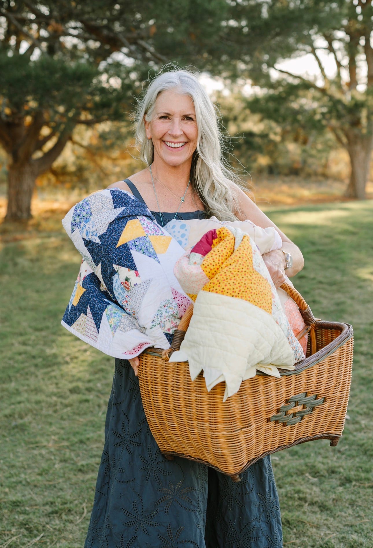 Allison Eyler stands outdoors holding a large wicker basket overflowing with colorful vintage and antique quilts, smiling in a teal eyelet dress with trees in the background.