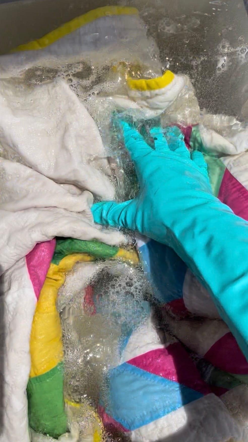 A gloved hand works a colorful quilt through soapy water during Allison Eyler's multi-step Cozy Clean restoration treatment for vintage quilts.