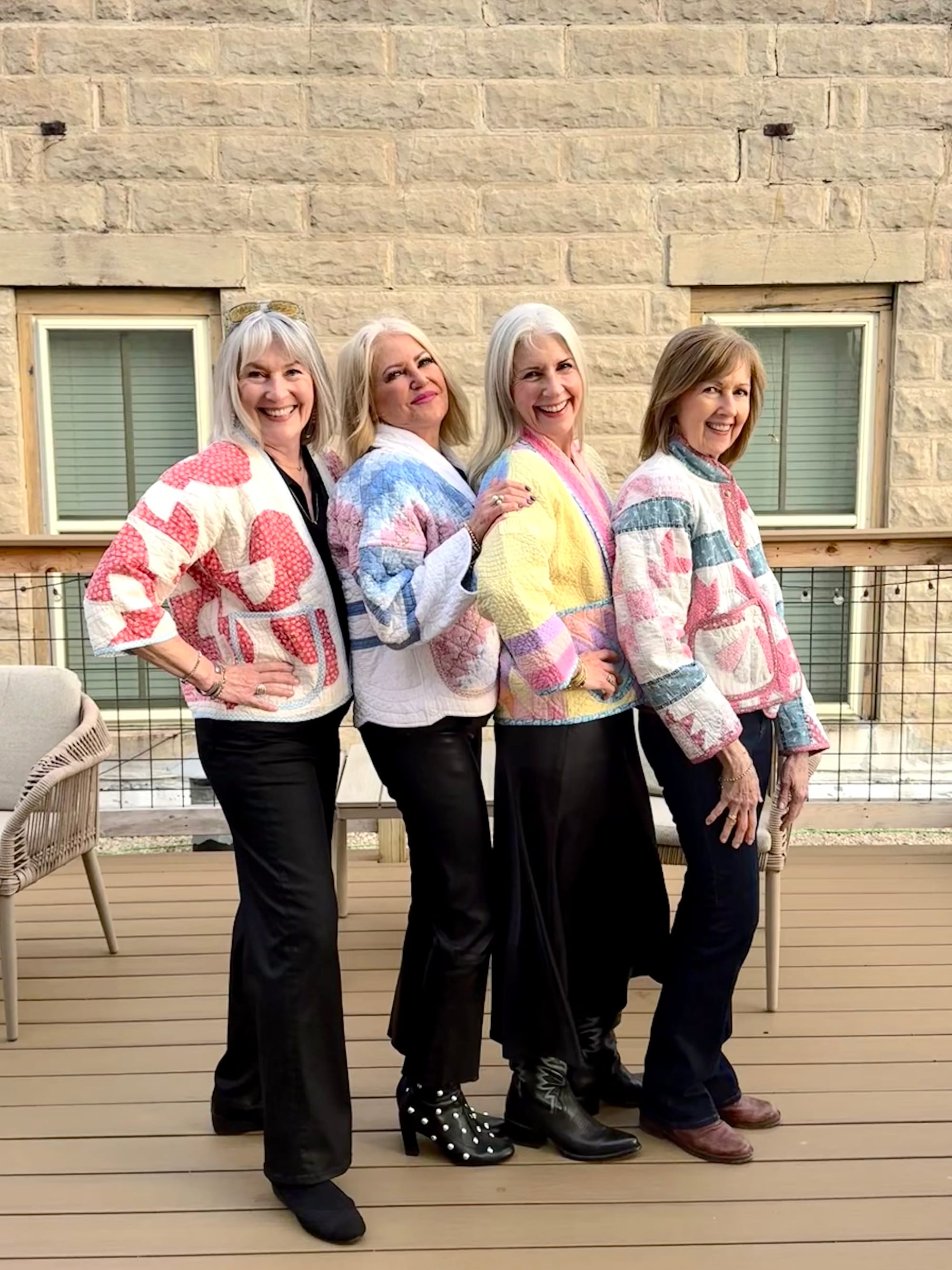 Four women stand on a deck wearing coordinating quilt jackets and coats made from upcycled vintage quilts in shades of pink, blue, yellow, and pastel patchwork.