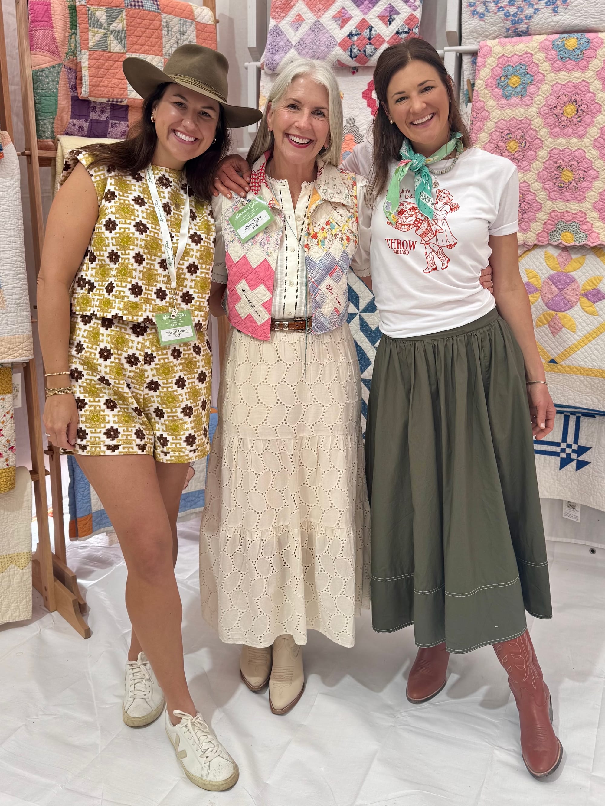 Allison Eyler stands between two smiling women at a quilt market or vendor show, wearing a patchwork quilt vest, with rows of vintage quilts displayed on wooden racks behind them.