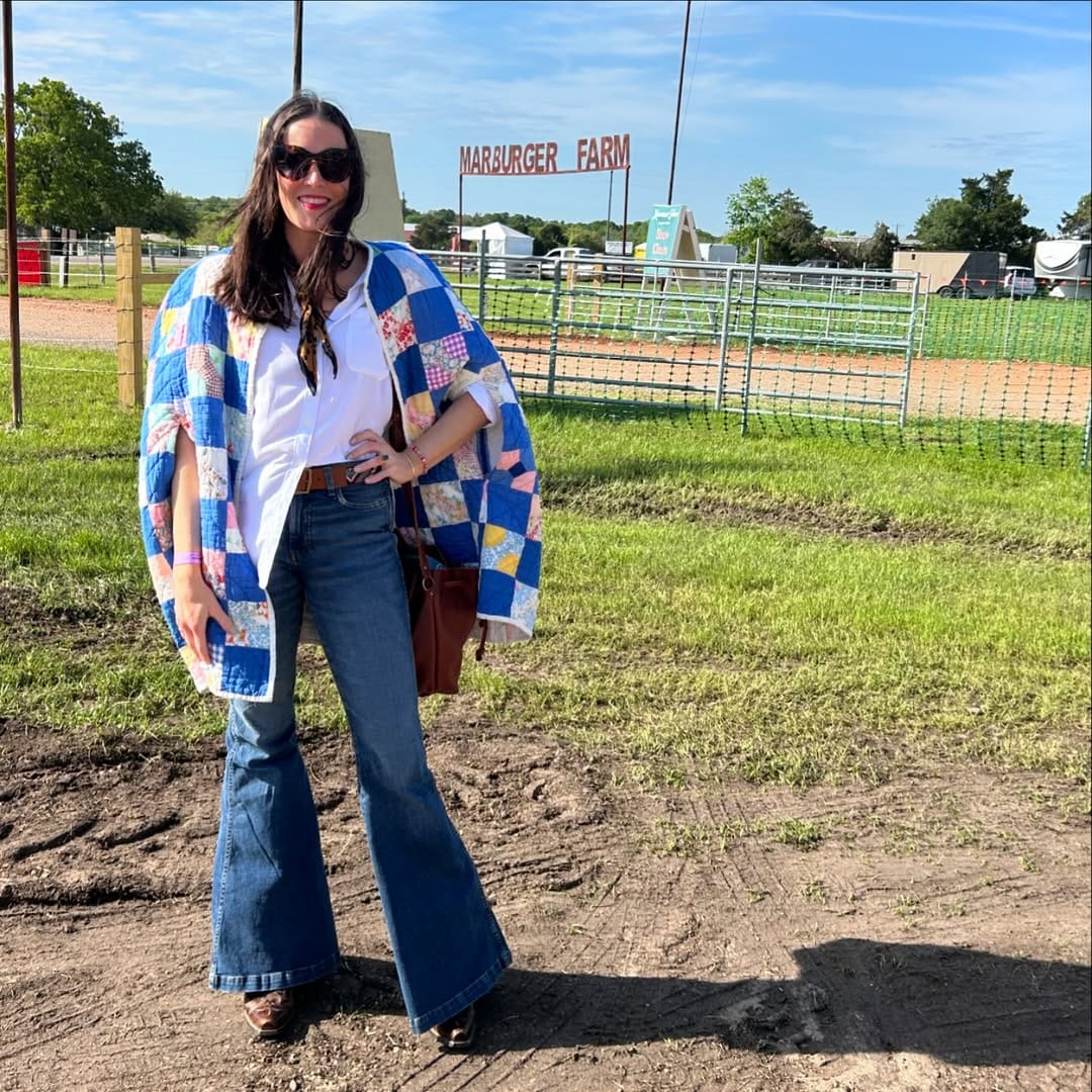 Sewist Lottie Dal poses at an outdoor market wearing a patchwork quilt coat made from vintage quilts over jeans and cowboy boots, with a Marburger Farm sign visible behind her.