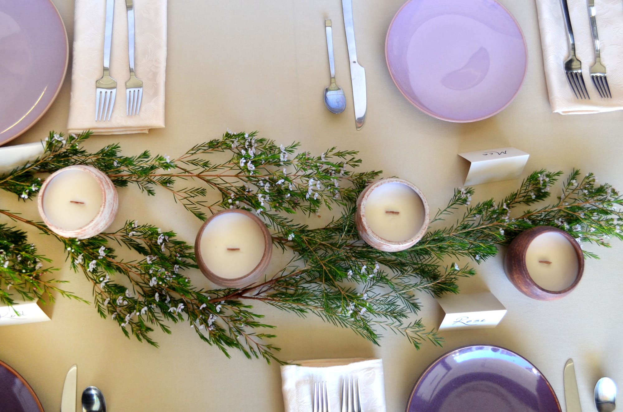 A dining table setting with pale purple plates, silverware, candles in wooden holders, and greenery with white flowers, featuring place cards for names.