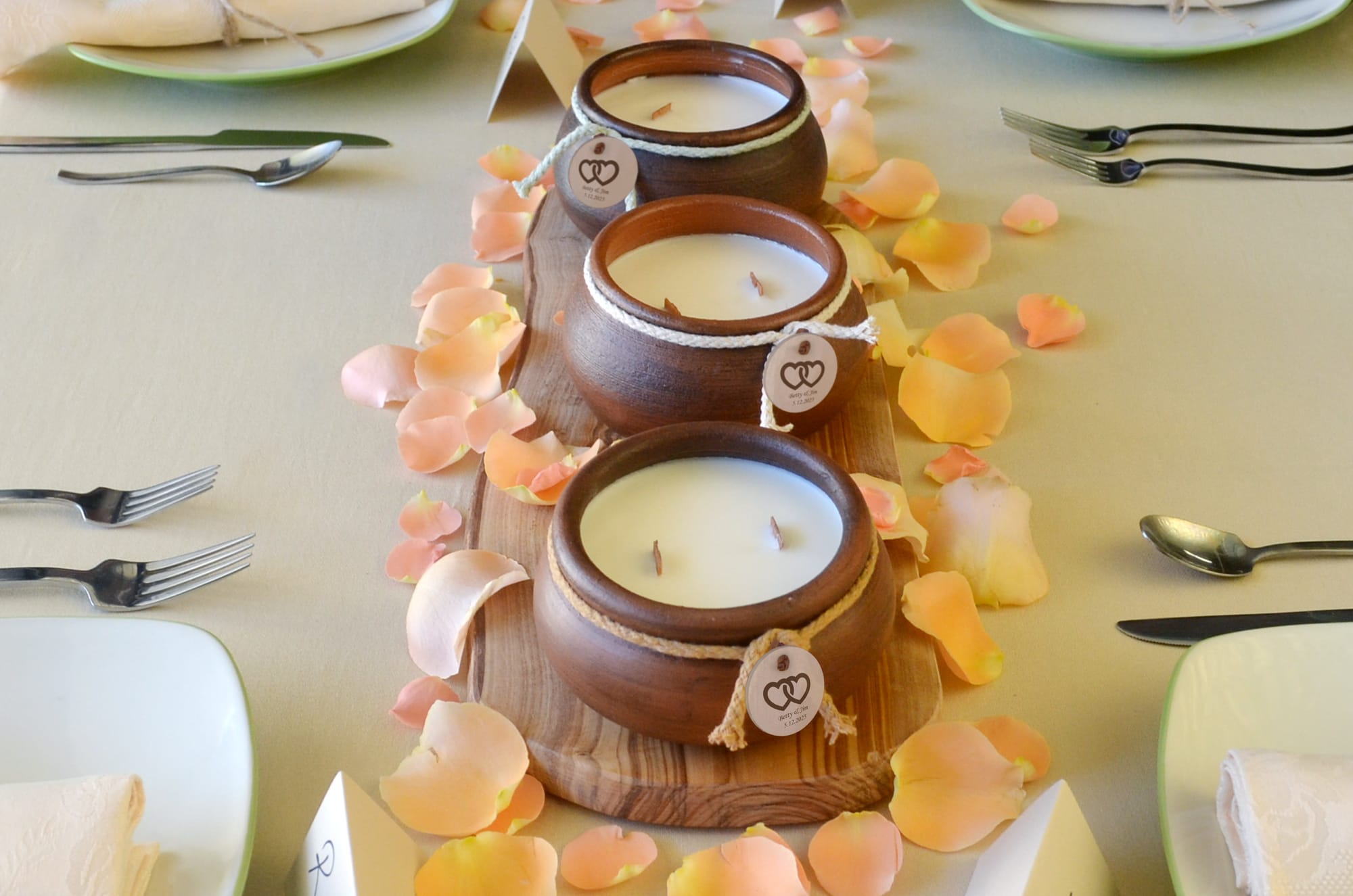 A decorative table setting with three brown ceramic candles on a wooden platter, surrounded by pale peach rose petals, and two settings with forks and plates.