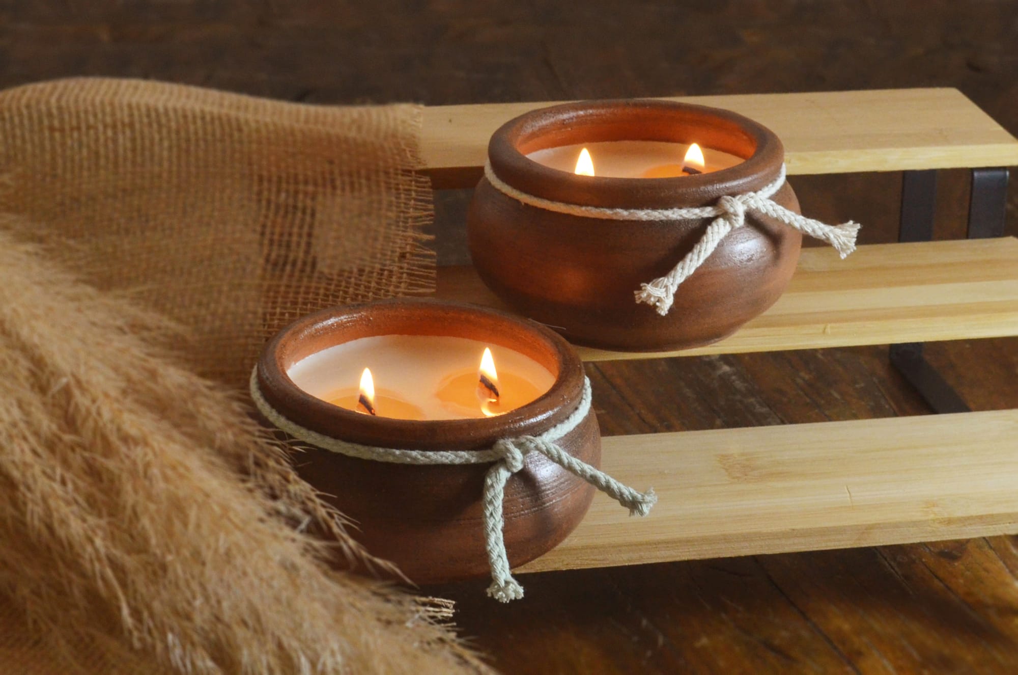 Two round terracotta candles with flickering flames, tied with white rope, sit on a light wooden shelf, surrounded by burlap and feathery foliage.