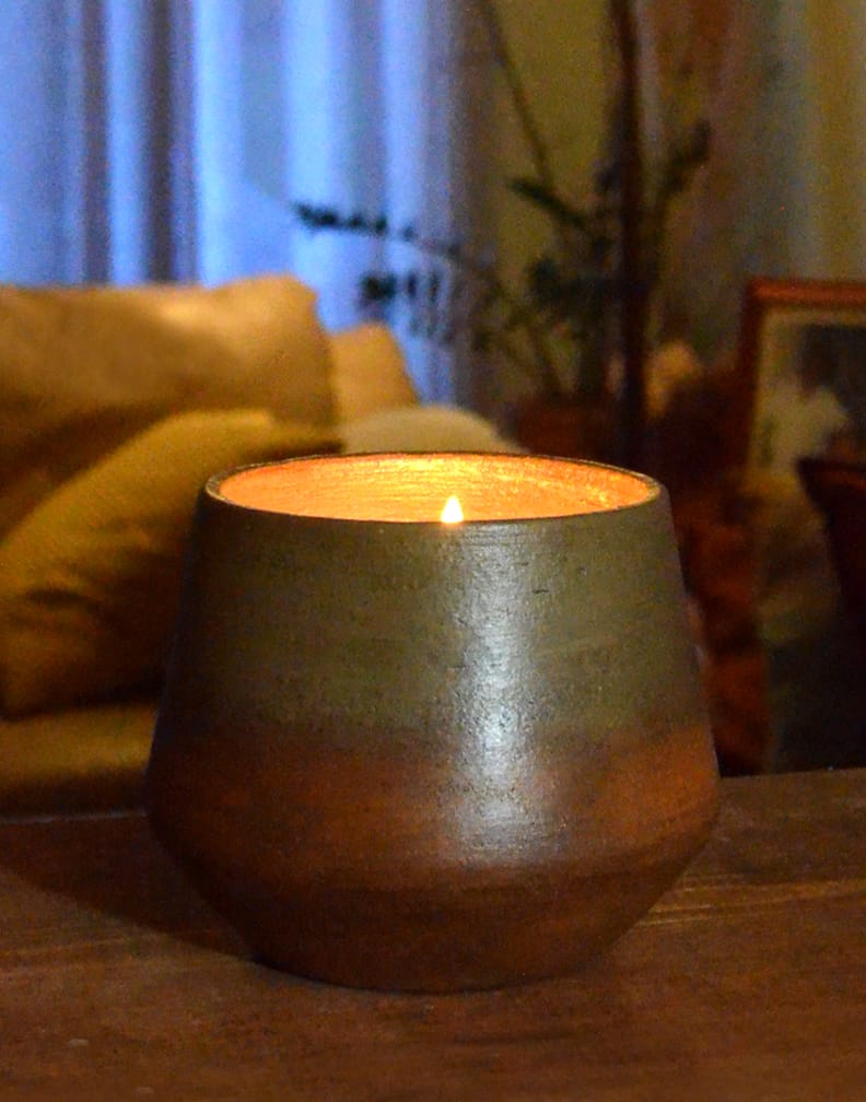 A round, brown candle holder with a flickering flame sits on a wooden table, with a blurred background showing a beige sofa and greenery.