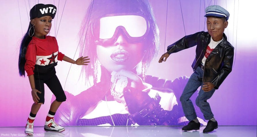 Two detailed marionette puppets modeled after Missy Elliott and Pharrell Williams pose in front of a large purple-toned image of Missy Elliott during a music video set.