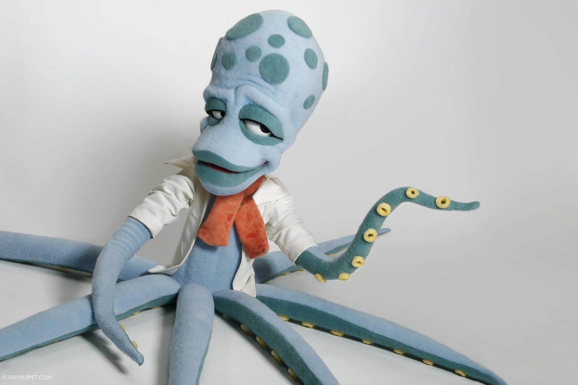 A blue octopus puppet with a spotted head and yellow suction cups wears a white lab coat and orange neck scarf against a plain light background.