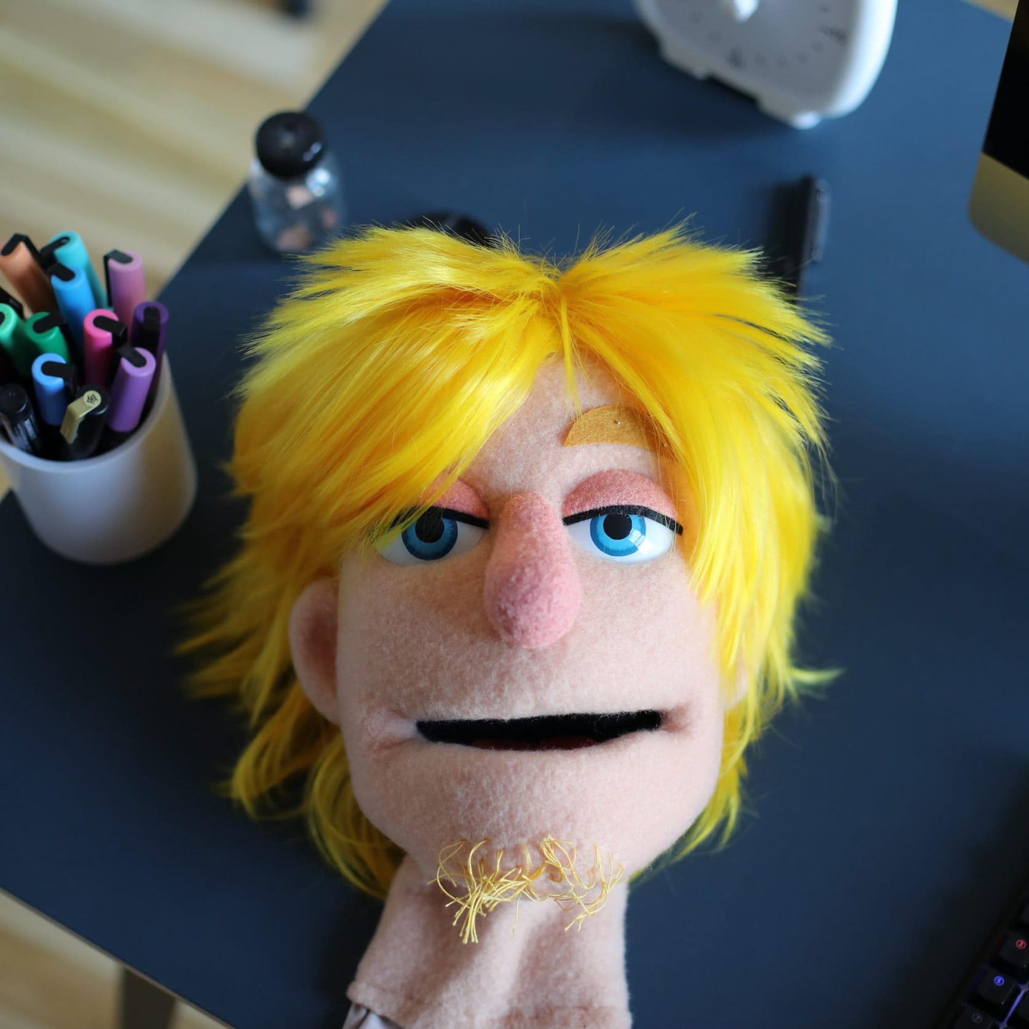 Close-up of a handmade puppet head with shaggy bright yellow hair, blue eyes, a pink nose and a small stitched mouth resting on a dark worktable beside cups of colorful markers.