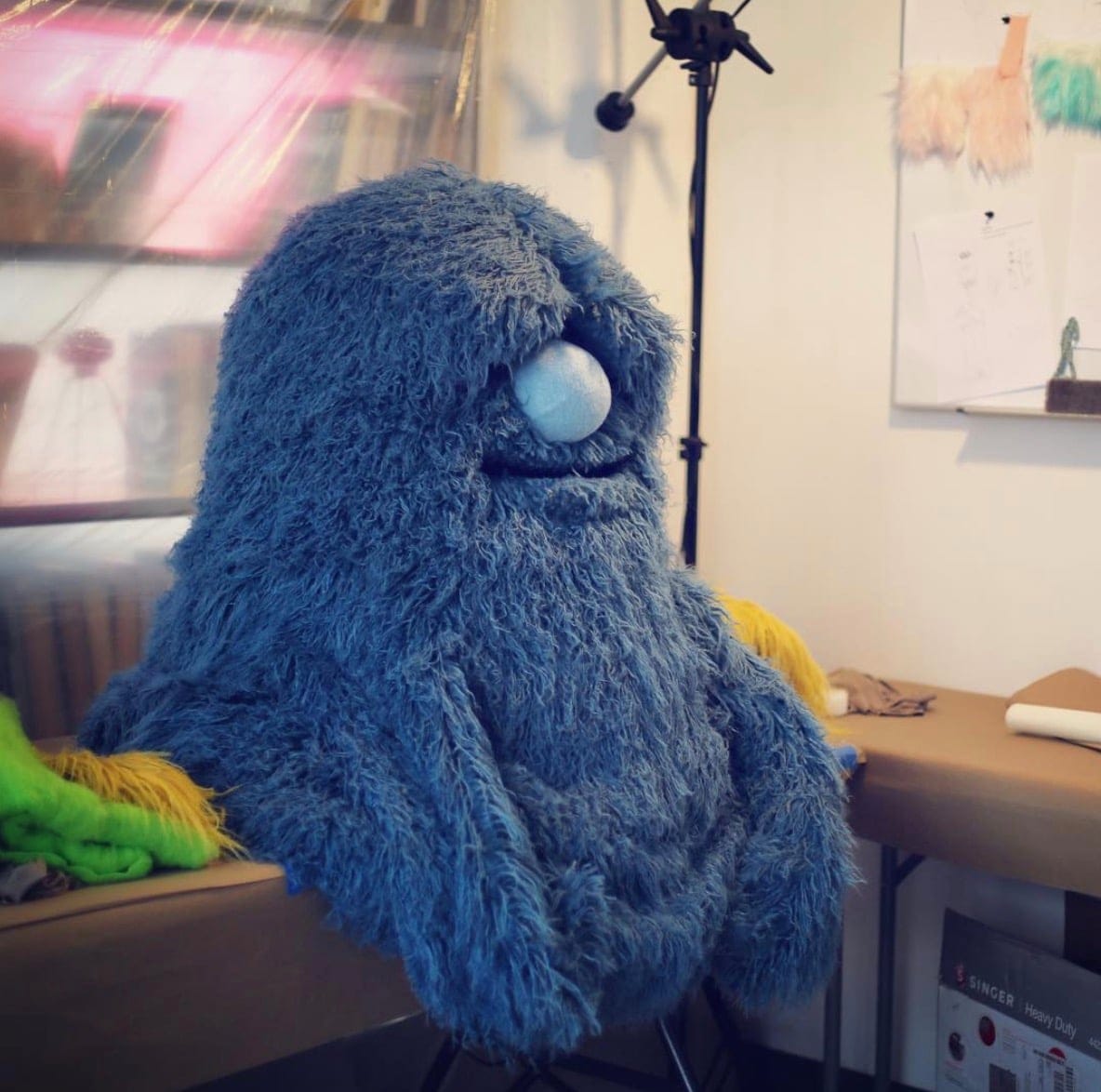 A large, shaggy blue monster puppet with a round light-blue nose sits in Furry Puppet Studio’s workshop beside worktables, fabric and a sewing machine.