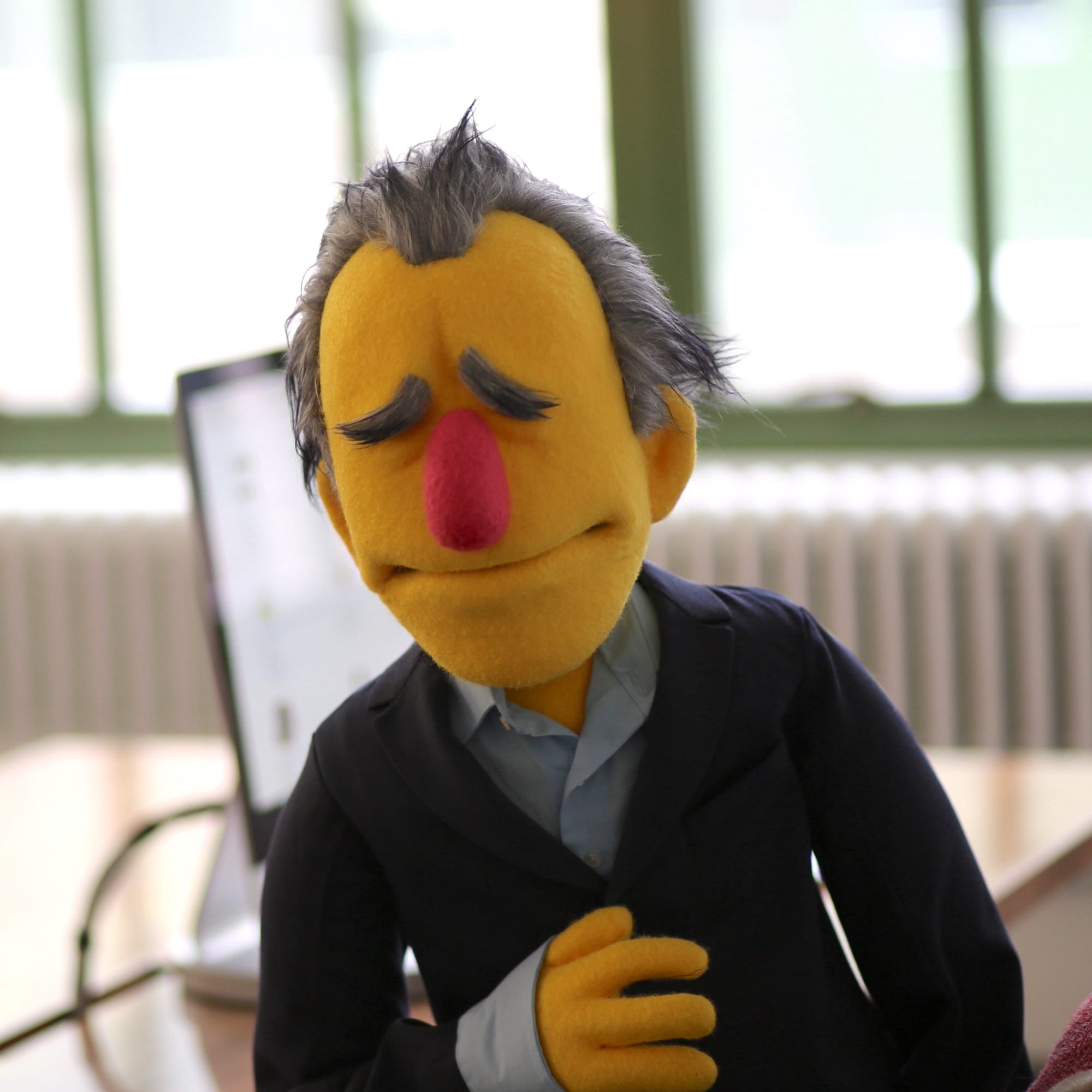 Close-up of a handmade puppet with yellow felt skin, gray hair and eyebrows, a pink nose, and closed eyes, dressed in a dark blazer and light blue collared shirt.