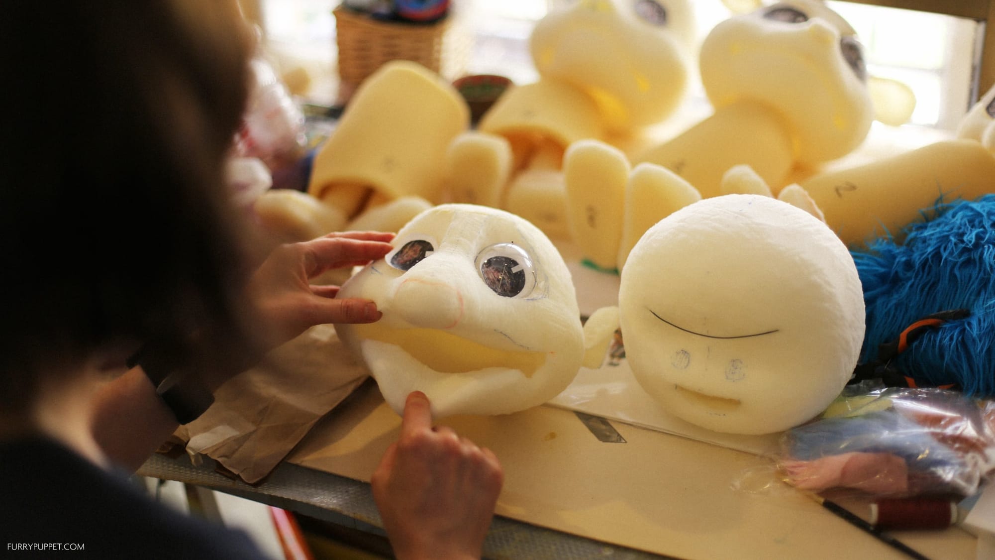 A maker adjusts the large eyes and mouth of an unfinished foam puppet head on a worktable beside another carved head and piles of yellow foam pieces.