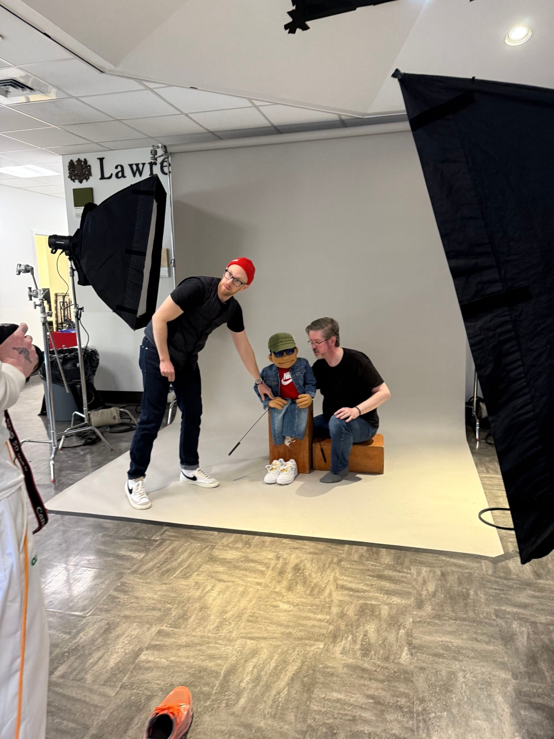 Two men pose a custom puppet dressed in a Nike jacket, cap and sneakers on a studio set surrounded by photography lights and equipment.
